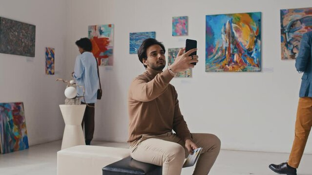 Medium full shot of young handsome Middle Eastern man in smart casual clothes sitting in art gallery during modern art exhibition, using smartphone, then taking selfie in front of paintings