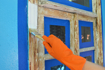 A worker's hand in an orange glove paints a window white with a brush. Painting an old wooden frame using masking tape. Household repairs, construction.