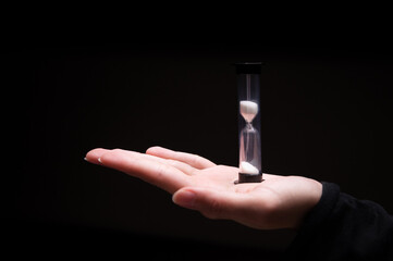 Close-up of a woman holding an hourglass in the dark. Fingers hold on both sides a plastic sand...
