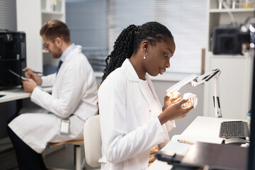 Side view of African American woman technician carefully looking at printed 3D bone sample using lamplight while sitting with defocused male coworker in laboratory