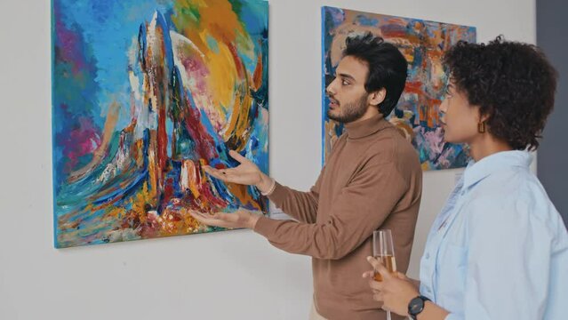 Medium shot of young Middle Eastern man attending modern art exhibition, standing in front of expressionist painting with female companion, explaining idea and composition, woman listening and nodding