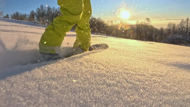 LOW ANGLE VIEW: Unrecognizable snowboarder guy riding powder, dog running next to him on sunny morning. Freeride snowboarder carving down the fresh snow at golden winter sunrise, dog running in snow