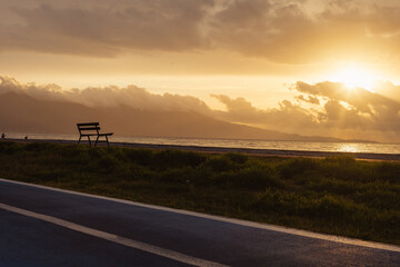 Dramatic cloudy sunset over shore. Empty bench, grass and blue bike line.