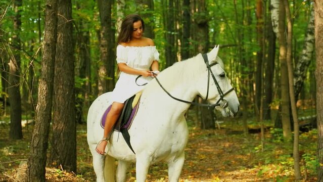 Young woman in white dress sits on white horse in park at autumn