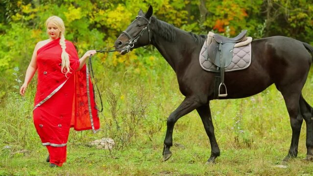 Blonde in red dress leads bay horse at autumn day in park