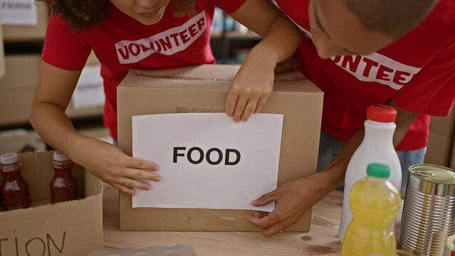 Beautiful young man and woman volunteers working together, putting food and paper in package to donate at community charity center
