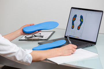 Female doctor orthopedist  holding custom made insoles in a clinic in front of the laptop with a test feet picture. - 718339920