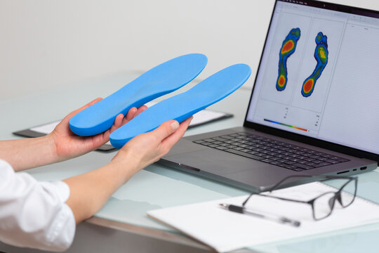 Female doctor orthopedist  holding custom made insoles in a clinic in front of the laptop with a test feet picture.