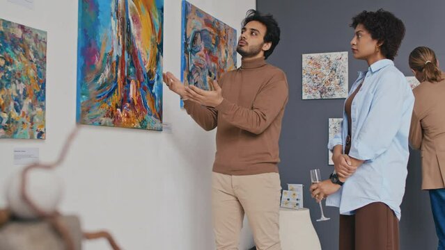 Medium shot of young male art critic standing in gallery at contemporary art exhibition, looking at modern artwork with female friend, talking about idea and technique, woman nodding in agreement
