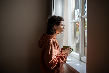 Lonely sad woman with cup of tea looking a window. Unsociable teenage girl spending weekend at home...