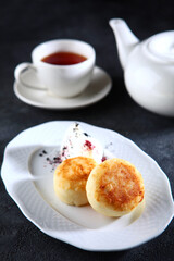 Cheesecakes on a white plate. Tea in a white porcelain teapot. Dark background. The concept of breakfast.