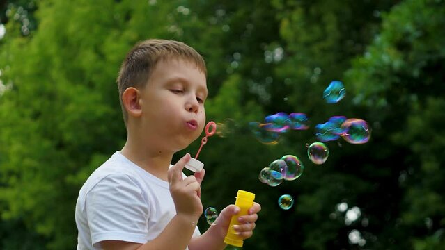Preschooler boy spends time blowing soap bubbles creating playful atmosphere slow motion. Playing in summer city park cheerful boy happily blows bubbles. Young boy delights in blowing soap bubbles