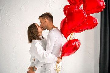 A girl kisses a guy on Valentine's Day holding a bunch of red foil balloons in the shape of a heart