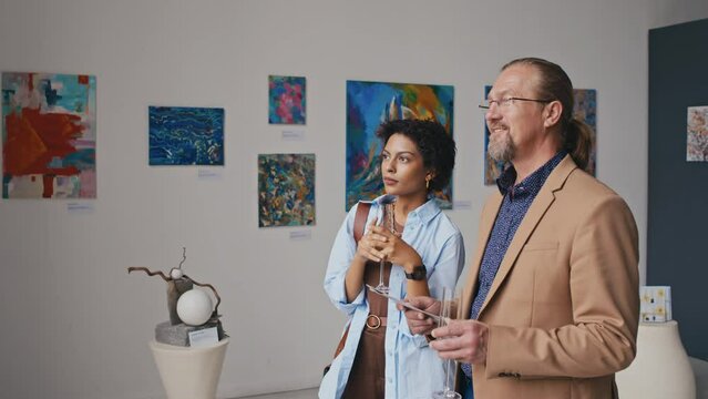 Medium shot of mature casually dressed Caucasian man and African American woman attending contemporary art exhibition at museum, viewing artworks, discussing paintings and sipping champagne