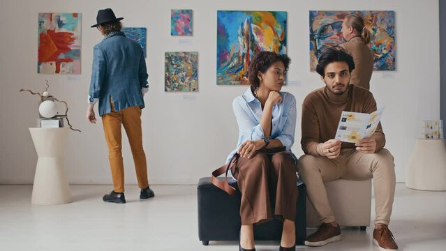 Full shot of young African American woman and Middle Eastern man sitting together on pouffe at modern art exhibition, reading booklet and discussing paintings, visitors looking at artworks