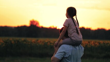 Daughter on shoulders of father gestures toward sunset. Daughter sits on shoulders with support of...