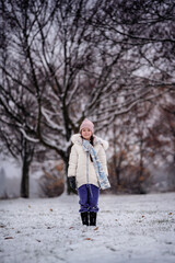 A young girl standing in a snowy landscape, with a backdrop of leafless trees, wearing a winter coat, a pink beanie, and a scarf