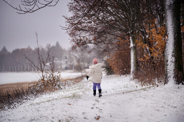 Fototapeta na wymiar The photo captures a child in a white coat and purple pants joyfully running in a snowy landscape, with snowflakes gently falling around.