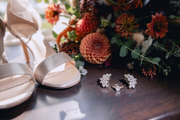 Obraz na płótnie Canvas Wedding accessories for the bride on her wedding day. A golden wedding ring with stones and earrings lie on a dark brown wooden table. A pair of milky wedding sandals and a bouquet of garden flowers. 