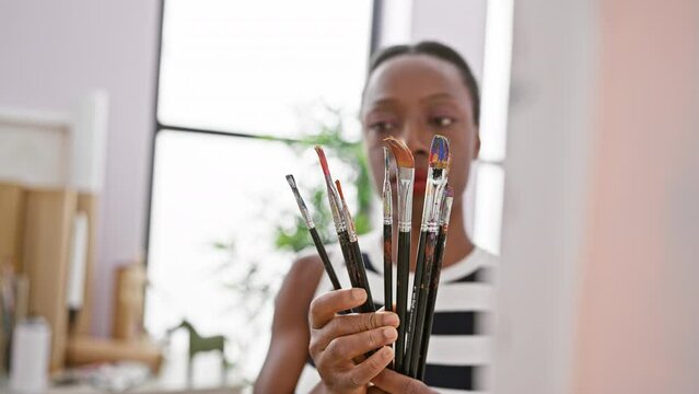At her art studio, the african american woman artist, engrossed and relaxed, makes a crucial decision, choosing the perfect paintbrush to breathe life into her canvas.