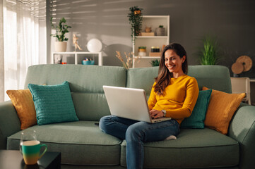 Portrait of a woman using laptop while sitting on a mint couch at home - obrazy, fototapety, plakaty