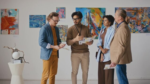 Full shot of confident Arab male art gallery employee talking to group of diverse visitors at modern art exhibition, answering questions, pointing at information in leaflet and paintings on display