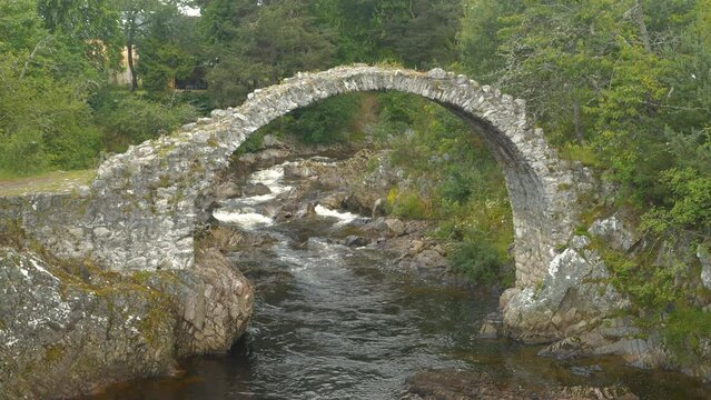 Mountain River Dulnain flowing under old rubble Carrbridge Packhorse Bridge. Lush greenery around beautiful remains of a stone arched bridge with an interesting history. Tourist attraction in Scotland