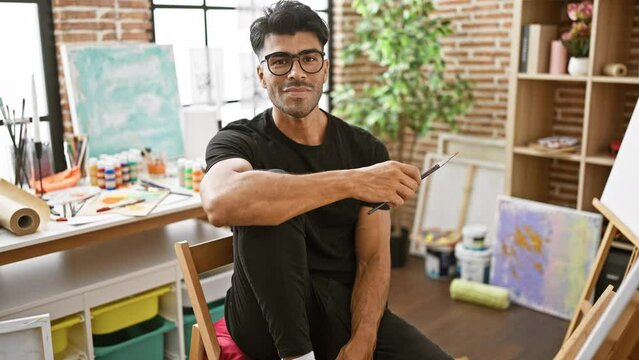 A handsome hispanic man with glasses and a beard holding a paintbrush in a bright art studio.