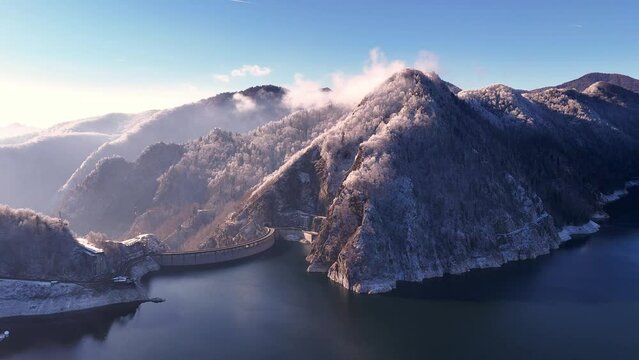 Aerial view of Water dam between snow covered mountains. Reservoir between high mountains. Hydroelectric power plant. Frozen trees in the mountain forest. Vidraru Reservoir in the Fagaras.  