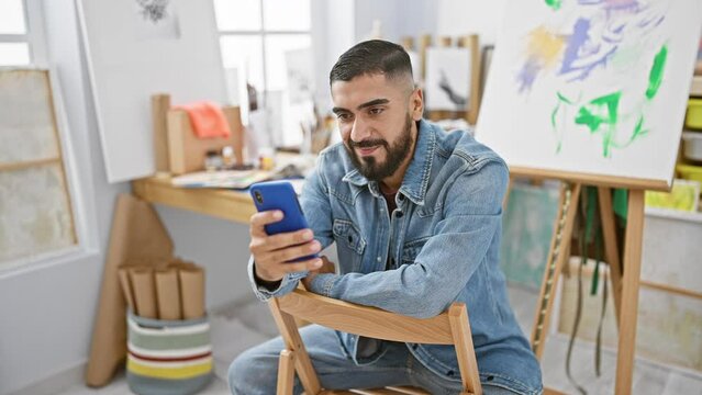 A young bearded man in a denim jacket using a smartphone in an art studio.
