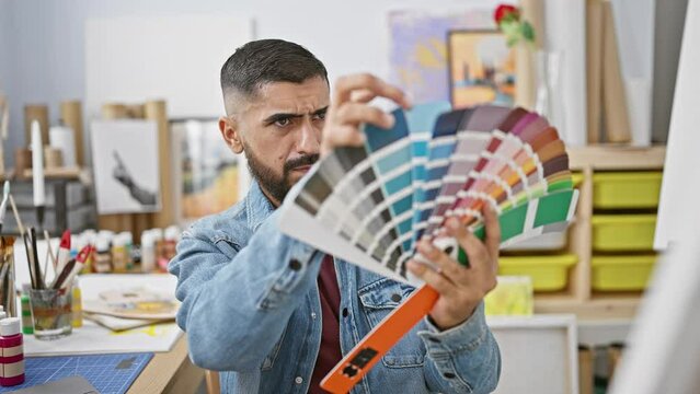 A focused man examines a color swatch in a creative studio, surrounded by art supplies and paintings.