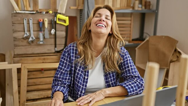 Crazy-cute, young beautiful latin carpentry worker puffing cheeks, amusing expression in the workshop. blowing air, hilarious face sits at table