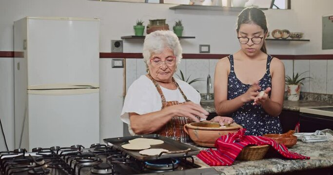 The Latina grandmother instructs her Latina granddaughter to make artisanal tortillas in her home kitchen.