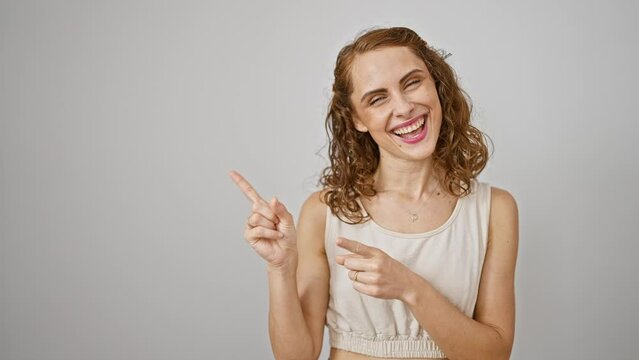 Confident young woman smiling, cheerfully pointing with both hands to the side, looking at the camera on an isolated white background, giving a welcoming gesture for product advertisement.
