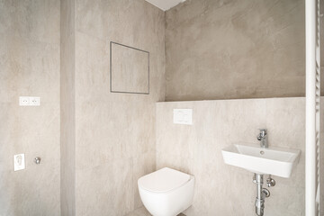 Small bathroom with modern beige tiling,wc and sink. Room is in process of construction or reconstruction,because part of wall is not covered with tiles,and therefore toilet is not yet completed. - obrazy, fototapety, plakaty