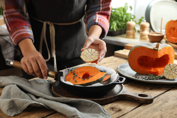 Woman adding thyme and garlic to pumpkin slices in frying pan at wooden table in kitchen, closeup