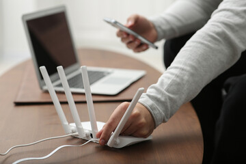 Man with smartphone and laptop connecting to internet via Wi-Fi router at wooden table, closeup