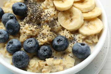 Tasty oatmeal with banana, blueberries and chia seeds served in bowl on white table, closeup