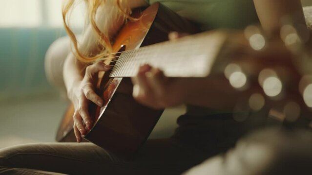 Cropped view of a red haired woman playing acoustic guitar while sitting on the floor, leaning back on sofa at home