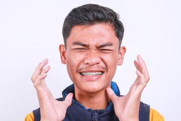 Close-up view of young Asian teenage guy with braces teeth raising hands and showing angry...