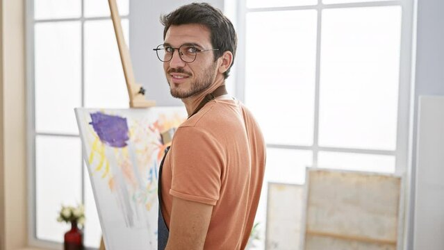 A bearded man painting on canvas in a bright studio wearing glasses and an apron, exuding creativity and focus.