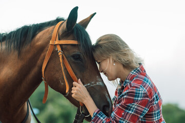 Obraz premium Happy blonde with horse in forest. Woman and a horse walking through the field during the day. Dressed in a plaid shirt and black leggings.
