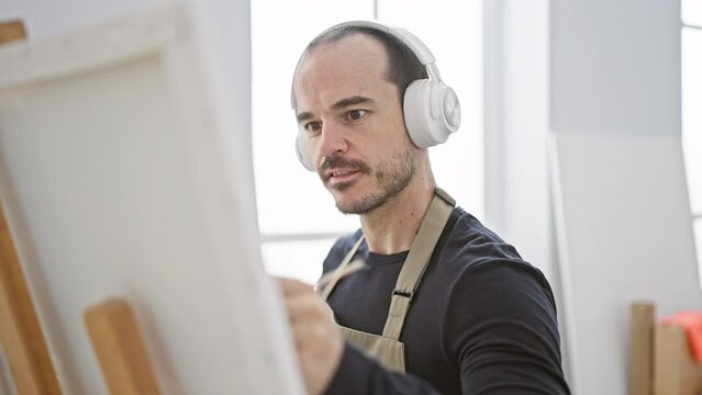 A focused bald man with a beard in headphones paints on a canvas at an art studio.