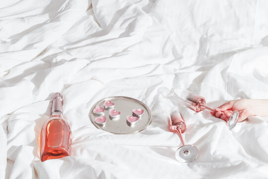 Pink champagne glass with metal shiny in female hand bottle of rose sparkling wine, candle hearts on bed. Natural daylight, star filter. Valentine's Day holiday, love concept, romance evening