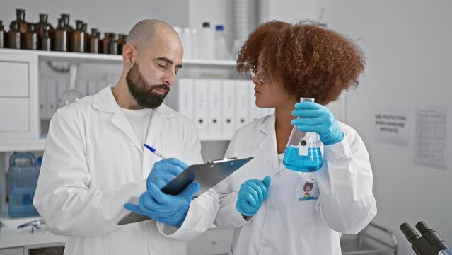Two serious scientists writing analysis results on clipboard, working together in a lab holding a test tube, amidst research documents and microscope