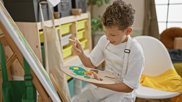 Adorable blond boy artist full of confidence, drawing his lovely idea with a radiant smile in indoor art studio, surrounded by canvas, paintbrushes & palette