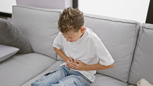 Cute little blond boy suffering from stomach ache, sitting on home sofa with a worried face, touching unsettled stomach