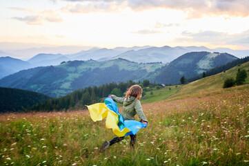 Happy girl running on hills with Ukrainian flag on shoulders. Back view of female child flutting Ukrainian flag. Little kid running in Carpathian mountains.