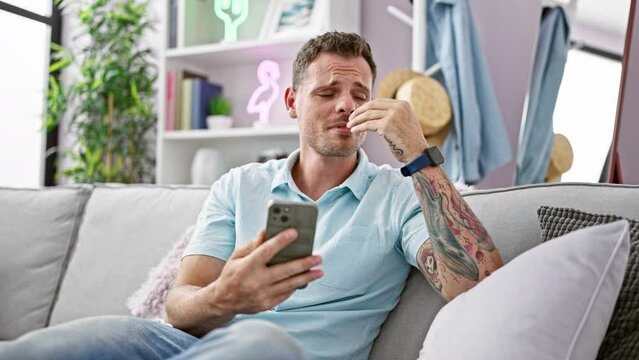A troubled man sitting indoors, emotionally reacting to content on his smartphone in a modern living room.
