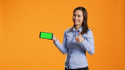 Confident girl shows greenscreen on phone display, standing over orange background and presenting...